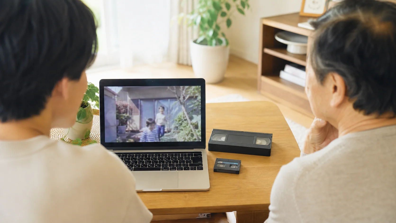 古いVHSと小型ビデオテープをそばに置き、親子でノートパソコンの家族映像を見返している様子
